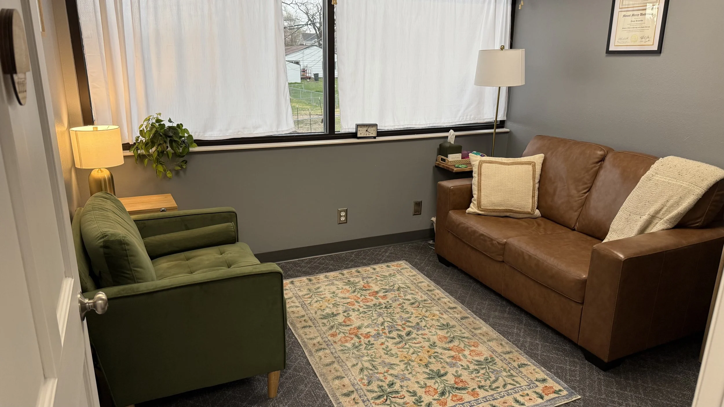 A small waiting room with a brown leather couch, a green armchair, a small wooden side table, and various lamps and decorations.