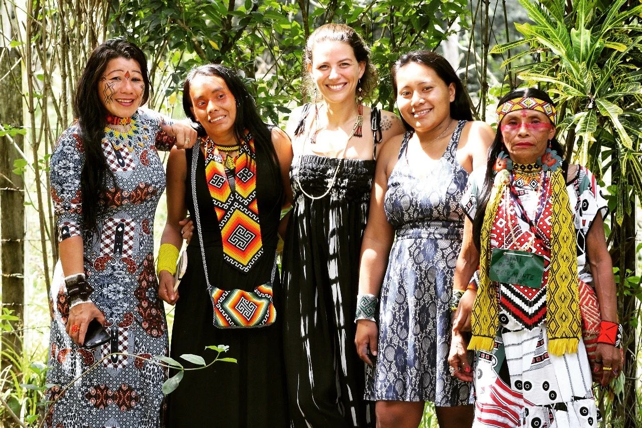 Cinco mulheres de pé, sorrindo, vestidas com roupas tradicionais e coloridas, em um ambiente ao ar livre cercado por vegetação.
