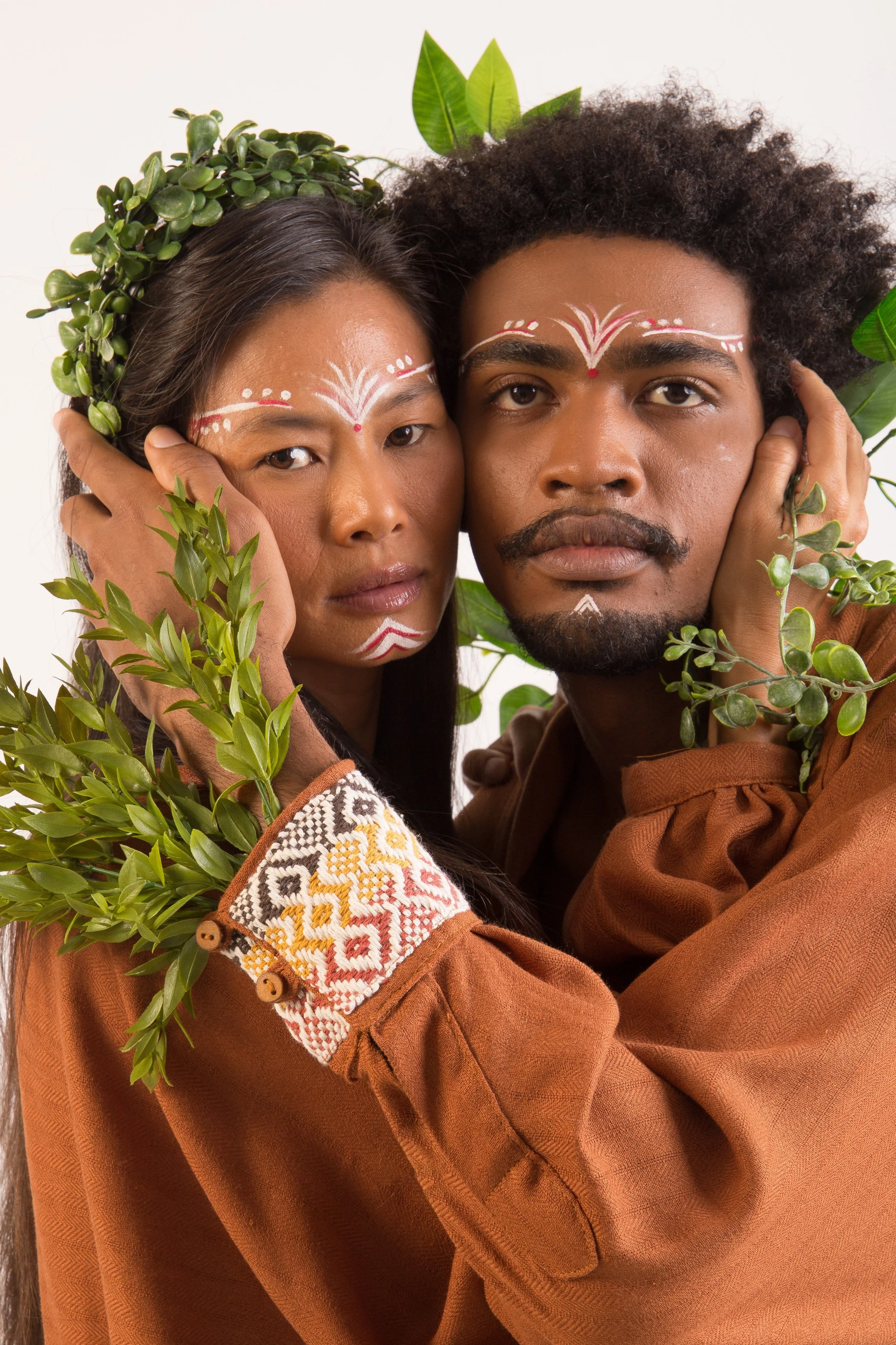 Casal com pintura facial tribal e vestes marrons, adornados com folhas verdes e cocar.