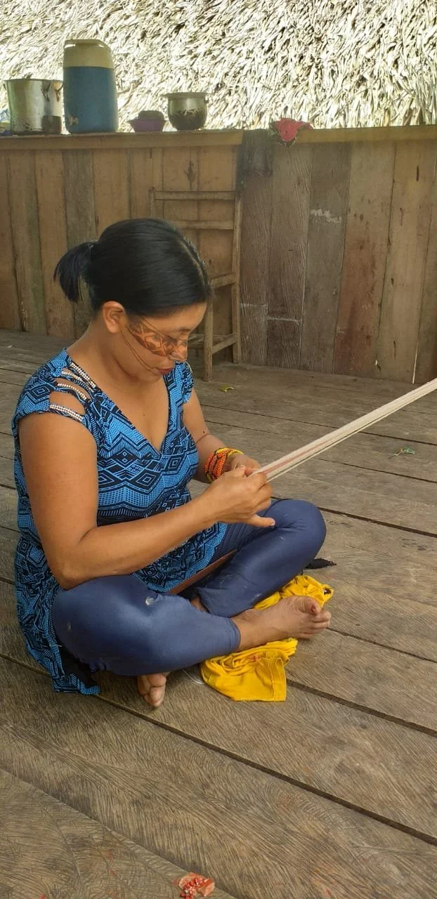 Mulher sentada no chão com roupas coloridas, usando uma ferramenta de artesanato em madeira, em ambiente de madeira com objetos ao fundo.