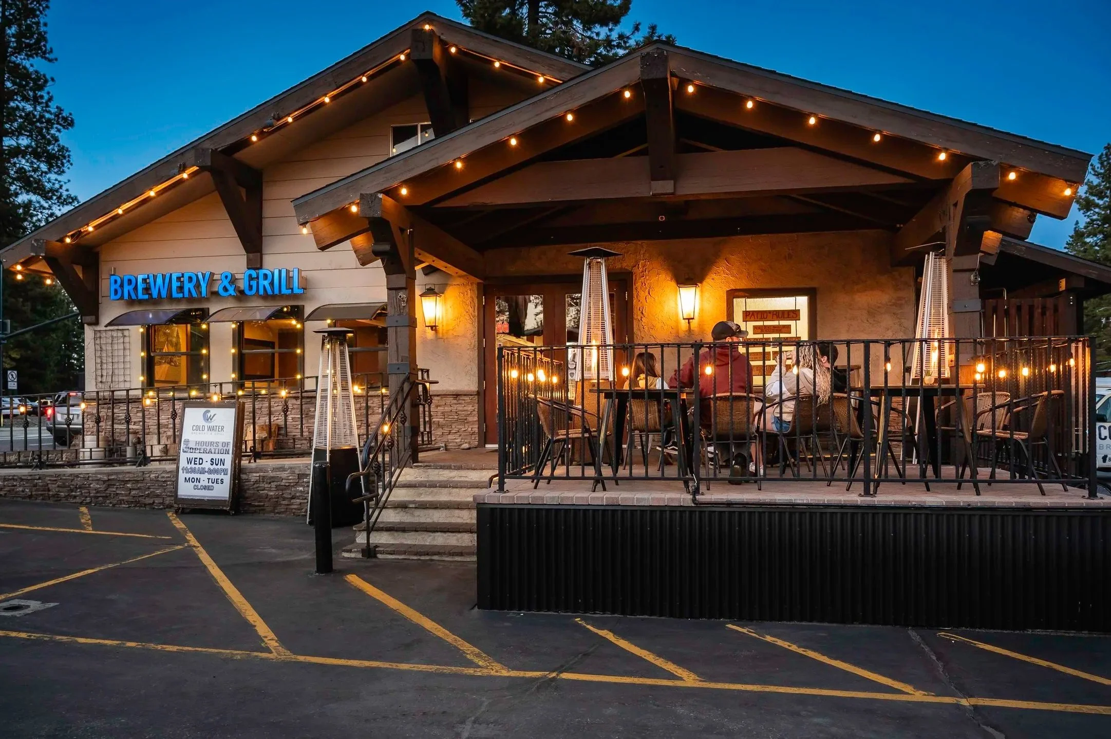 Exterior of a brewery and grill with outdoor seating, string lights, and people dining on a patio during evening.