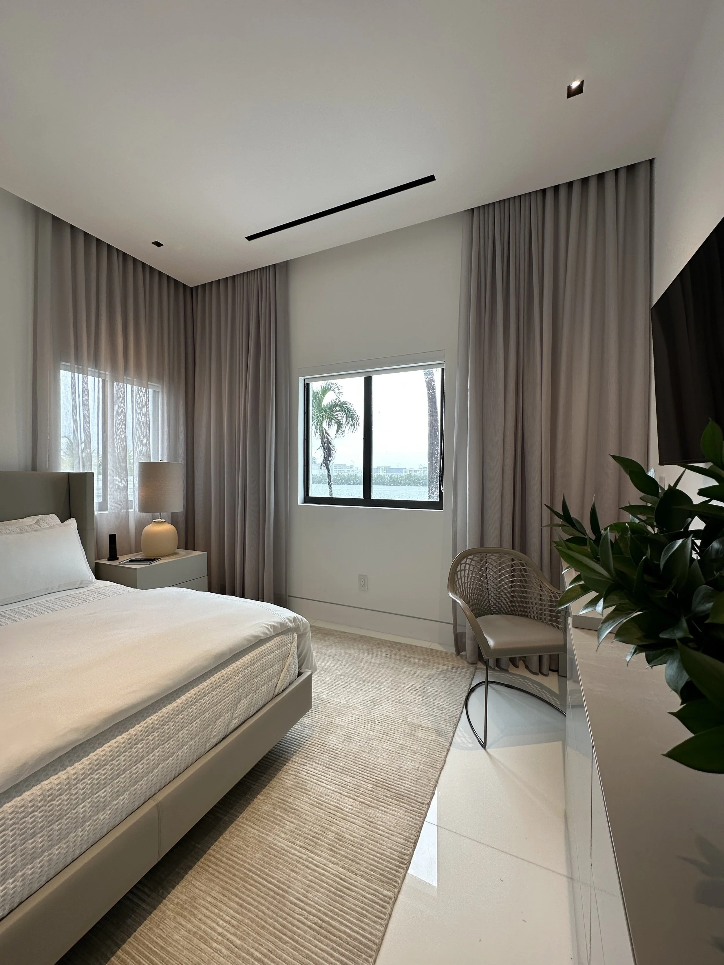 Modern bedroom with a large bed, white linens, bedside table with a lamp, large windows with curtains, wicker chair, indoor plant, and white glossy floor tiles.