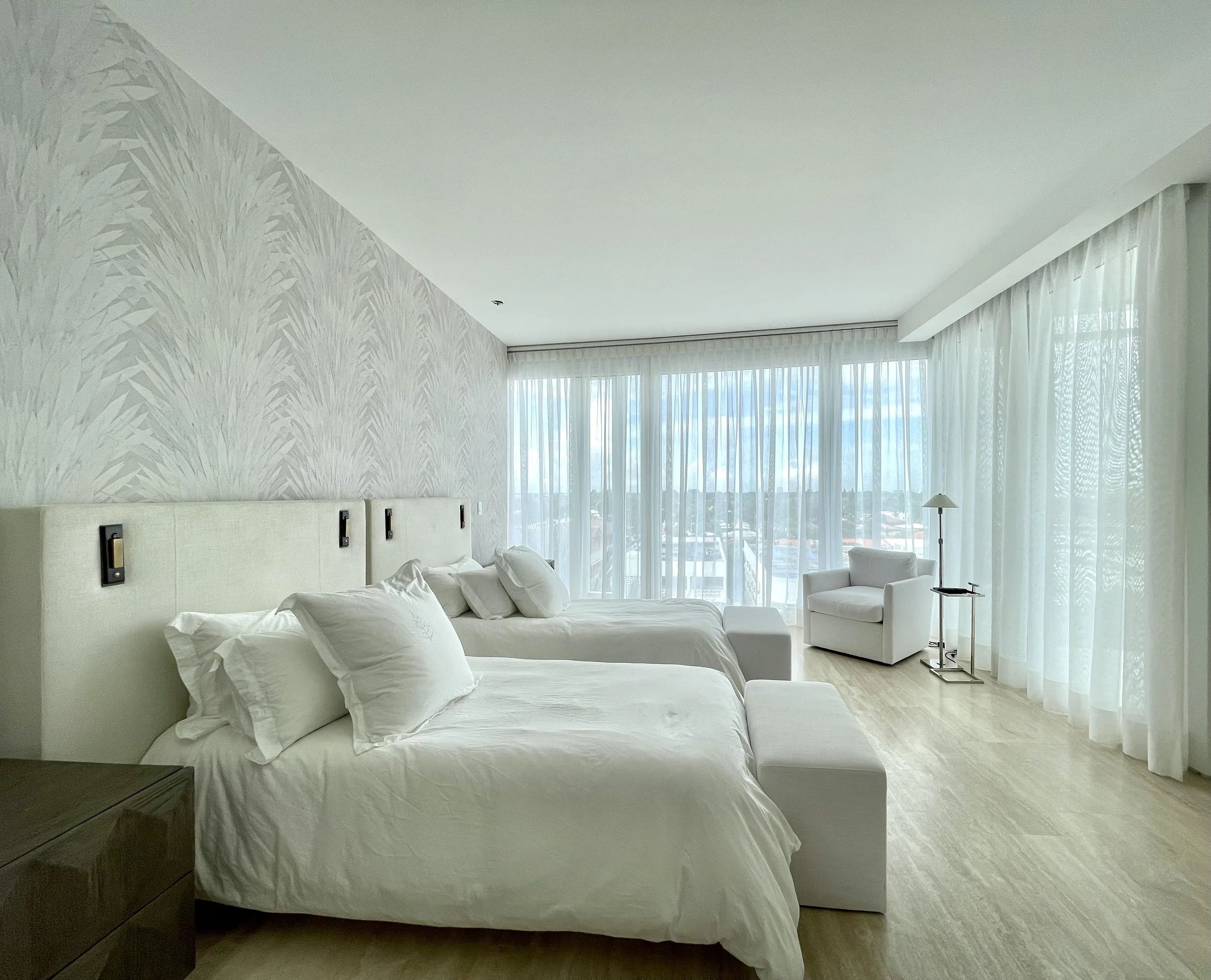 Modern hotel room with two white beds, decorative wallpaper, large windows with sheer curtains, a white armchair, and a side table.