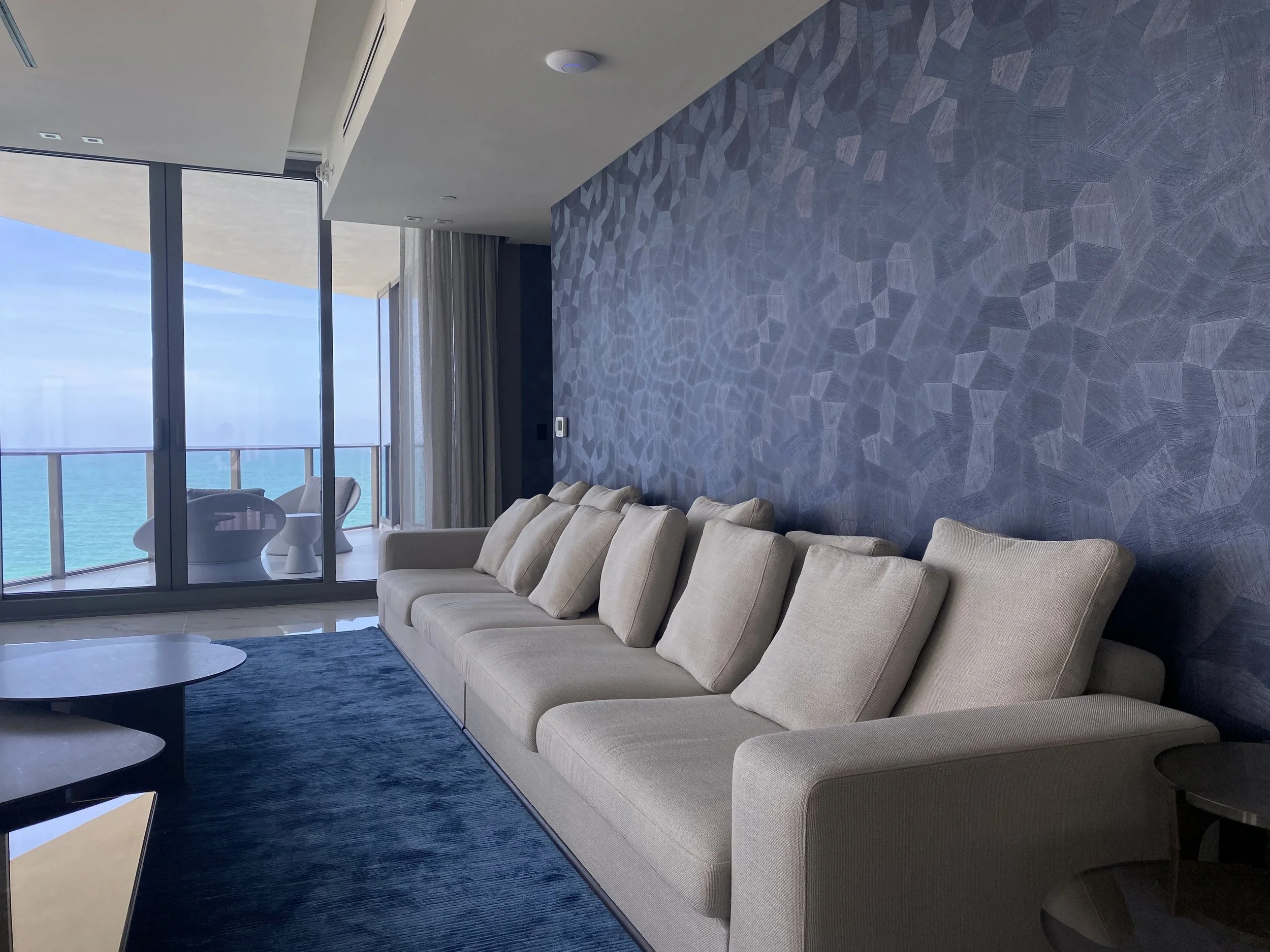 Modern living room with beige sofa, blue textured wallpaper, circular coffee tables, large glass windows, and ocean view balcony with chairs.