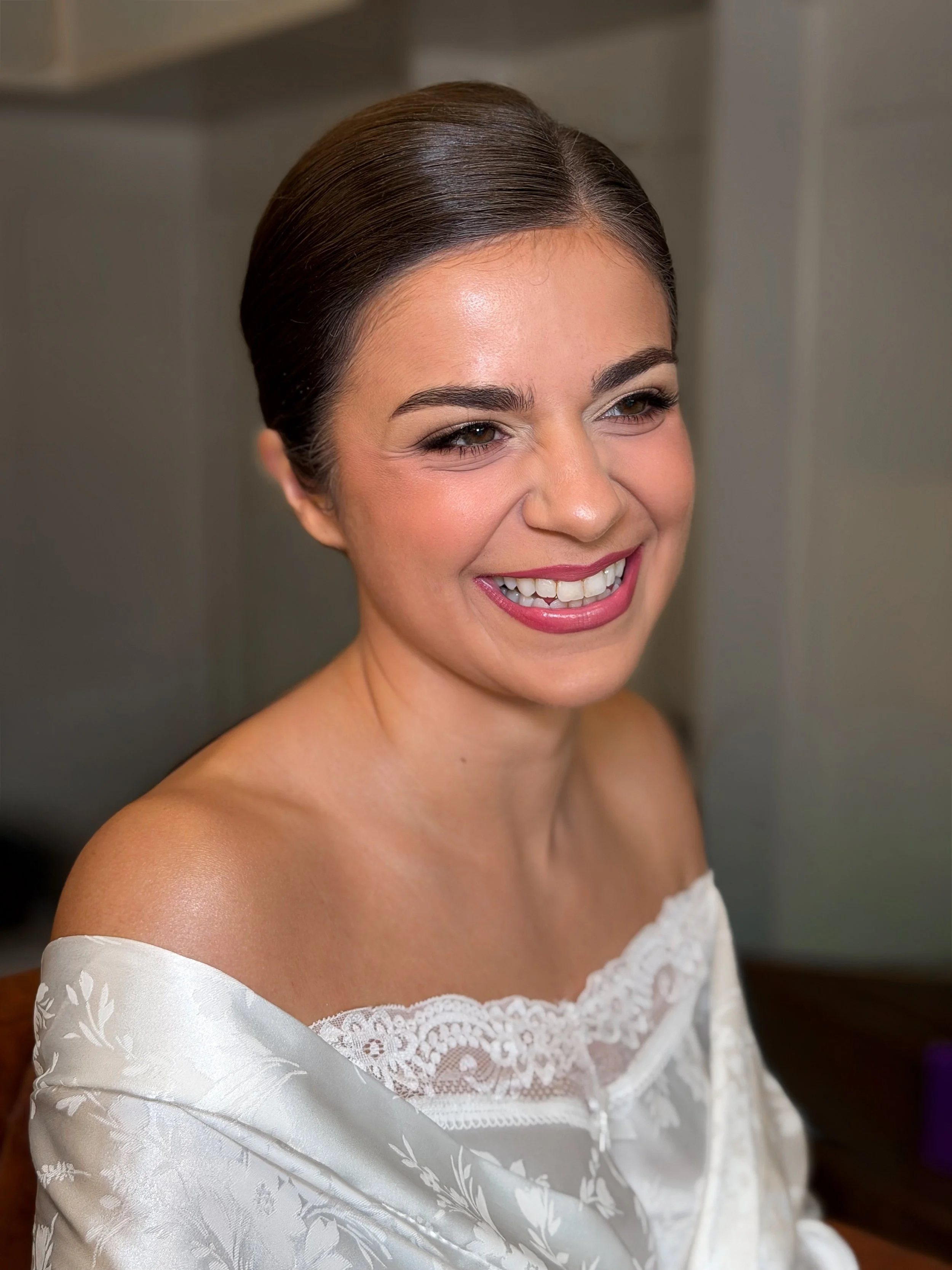 Smiling bride with natural makeup and slicked-back brown hair wearing a white off-shoulder lace dress.