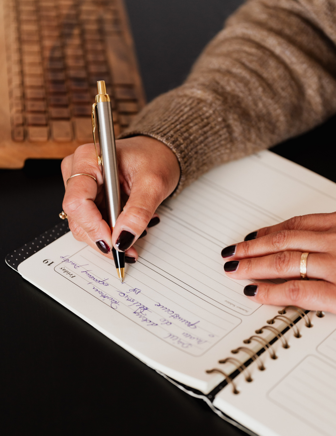 A person with dark nail polish writing in a planner with a black and gold pen. The person is wearing a brown knitted sweater and a gold ring. There is a wooden keyboard in the background on a black surface.