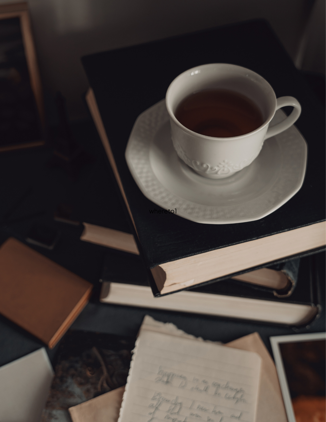 A white teacup filled with tea on a white saucer, placed on top of a closed black book, with other books and a torn piece of lined paper around.
