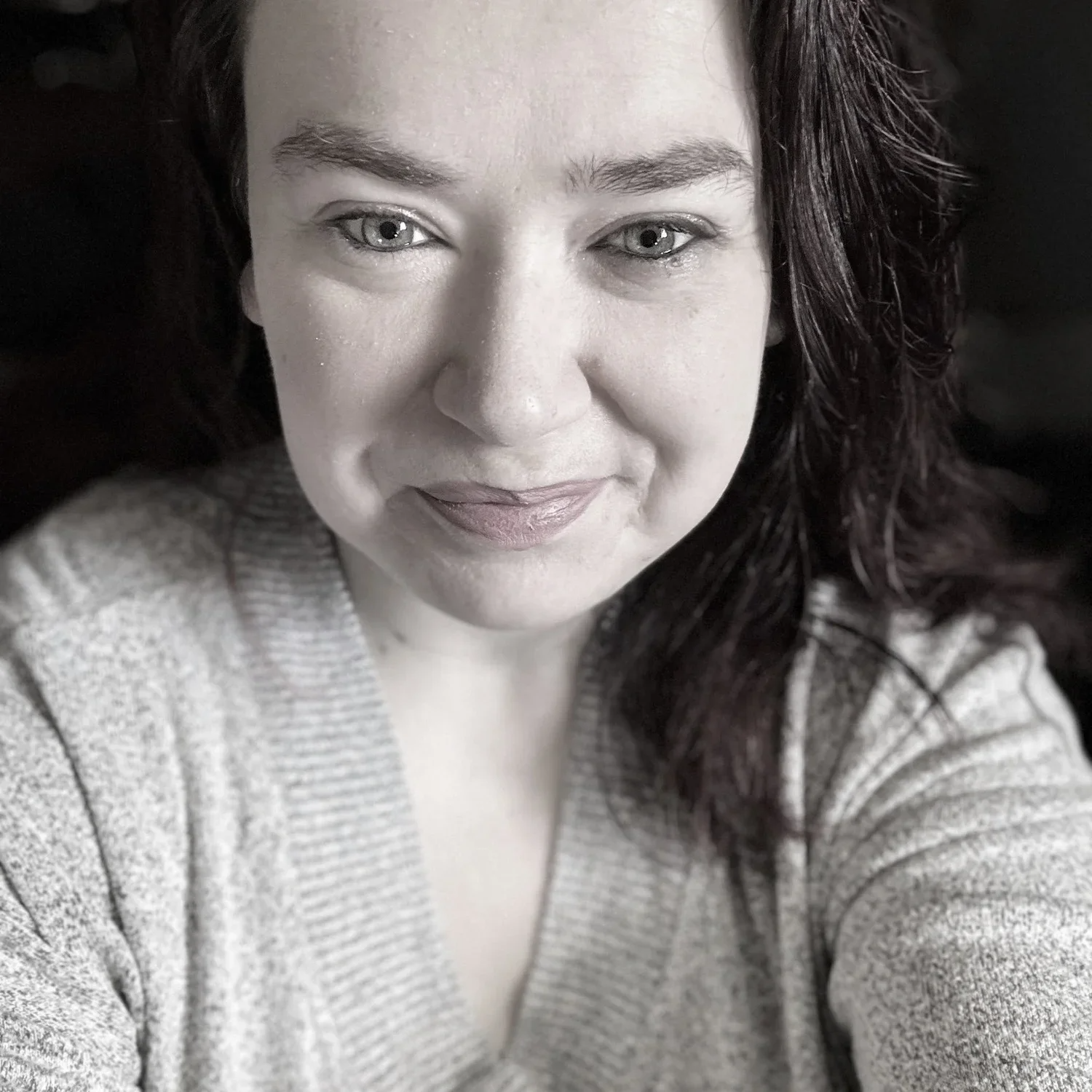 A woman with light skin, dark hair, and blue eyes is taking a selfie, wearing a light-colored sweater, and smiling slightly.