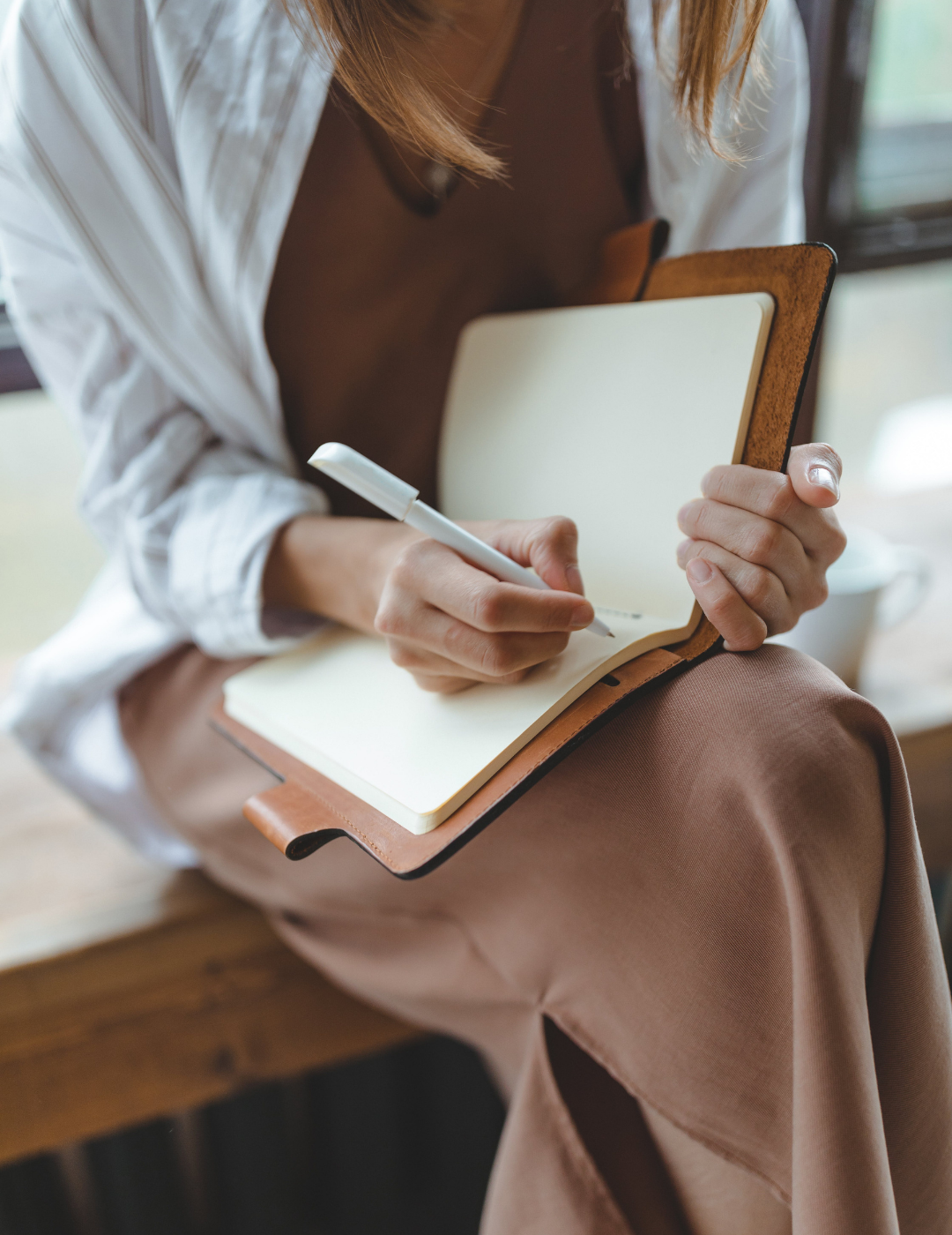 Person writing in a notebook with a pen while holding a closed laptop, seated indoors near a window.