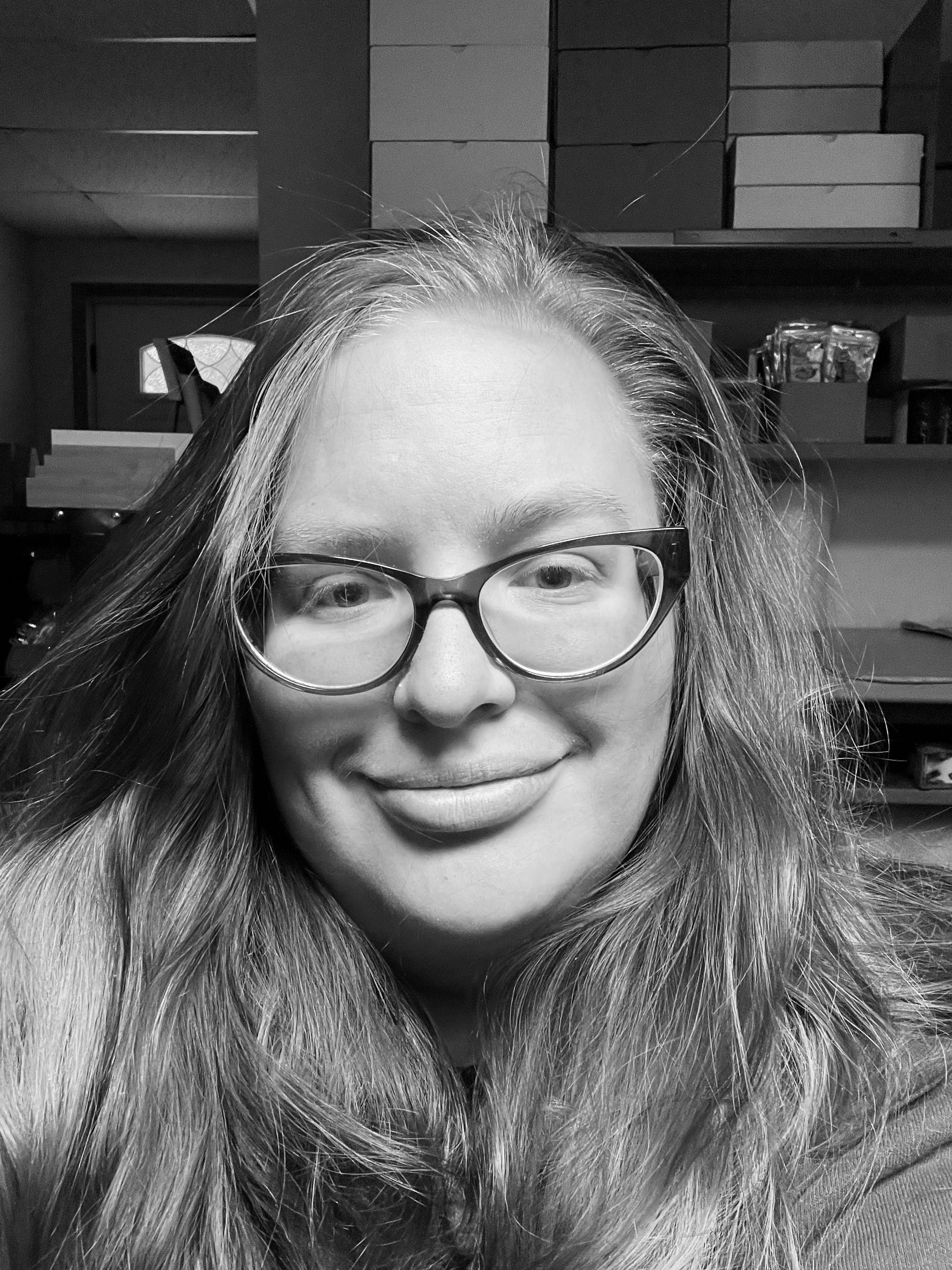 A woman with glasses and long hair smiling in a room with shelves of boxes and items in the background.