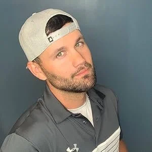 A man wearing a gray baseball cap backward, with a beard and mustache, looking at the camera. He has on a gray sports shirt with a white stripe, against a blue background.