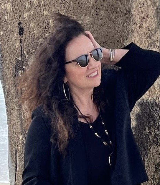 Woman with curly brown hair wearing sunglasses and hoop earrings, standing outdoors near a tree, smiling.