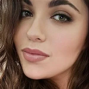 Close-up of a young woman's face with makeup, green eyes, and wavy brown hair.