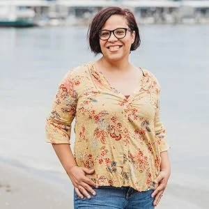 Woman with glasses smiling outdoors near a body of water.