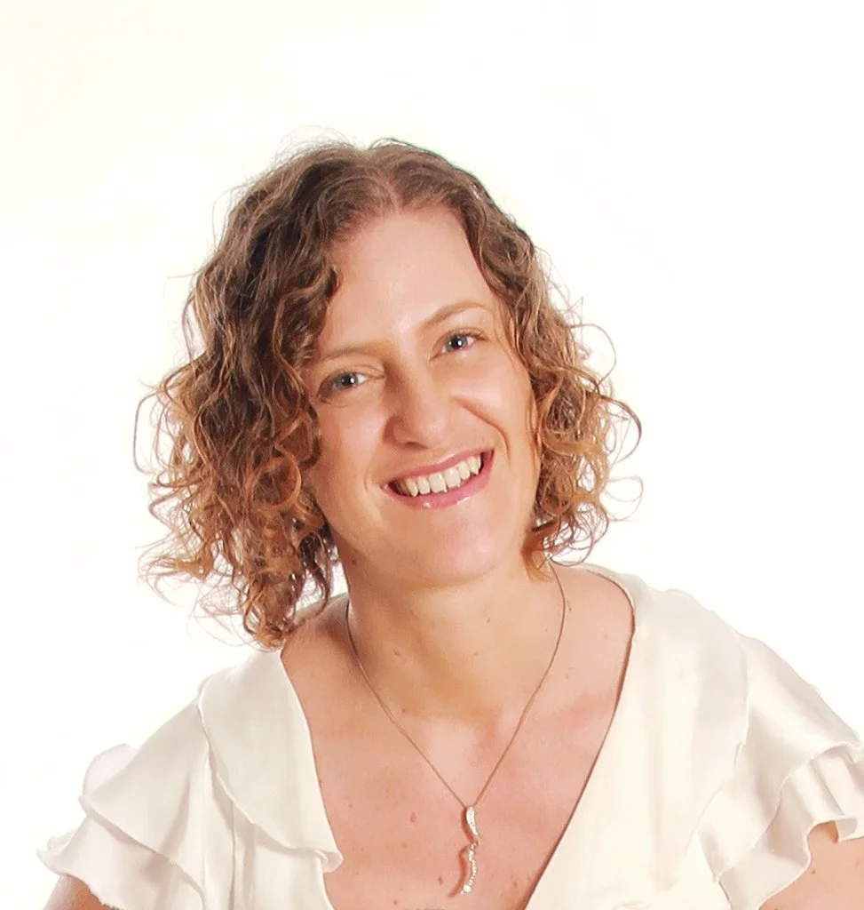Portrait of a smiling woman with curly reddish hair wearing a white ruffled top and a gold necklace with a snake-shaped pendant.