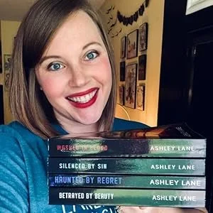 A woman with shoulder-length brown hair, blue eyes, and red lipstick holding a stack of five books by Ashley Lane, smiling at the camera in a room with framed pictures on the wall.