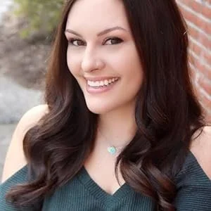 A young woman with long dark hair smiling outdoors next to a brick wall.