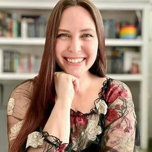 A woman with long brown hair smiling and resting her chin on her hand, indoors with a bookshelf in the background.