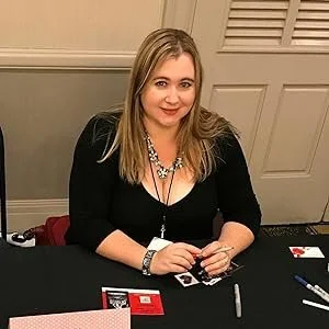 Woman with blonde hair wearing a black top and a necklace, sitting at a table with playing cards and markers.