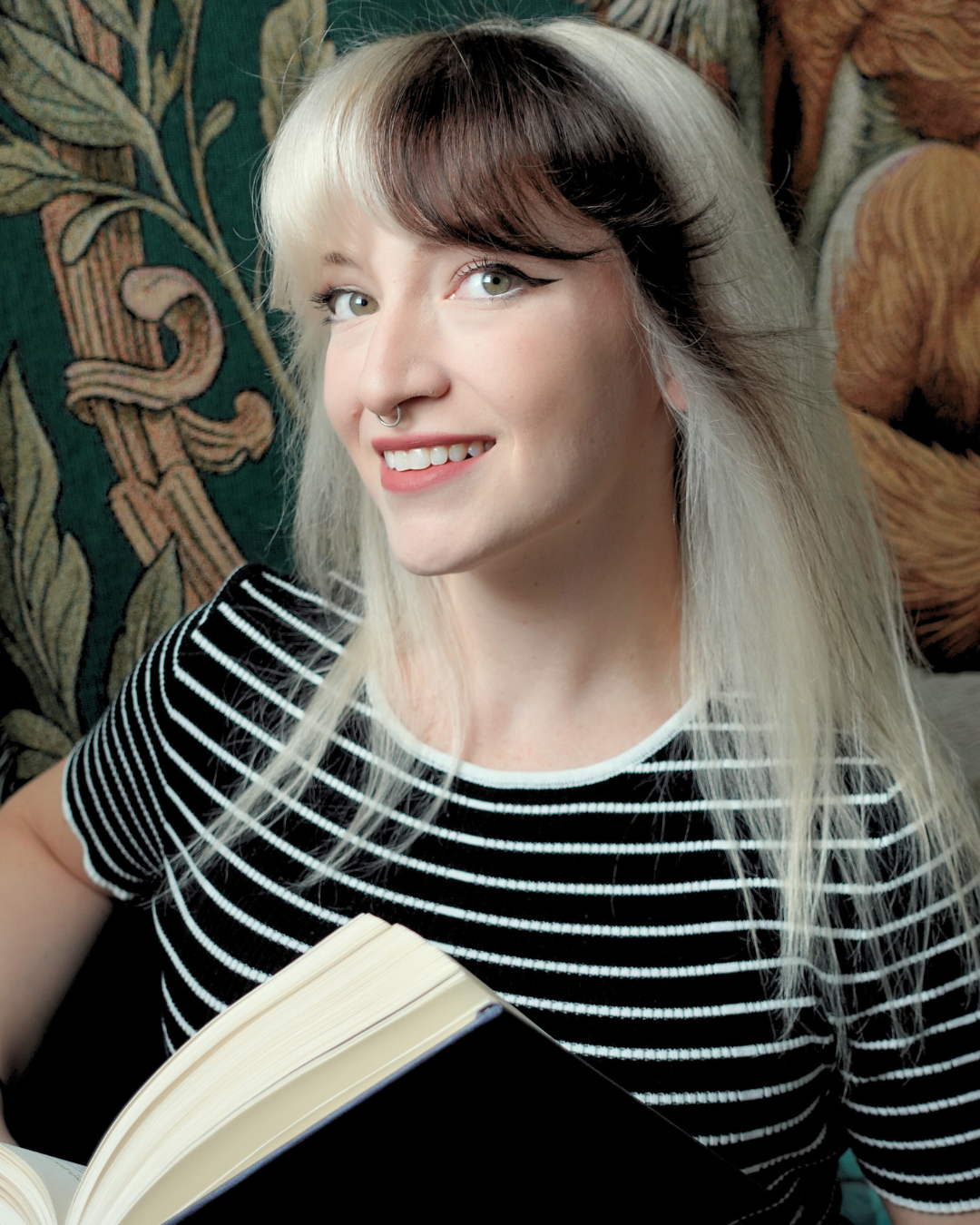 A young woman with blonde and brunette hair, wearing a black and white striped shirt, smiling and holding a book, sitting on a sofa with a patterned fabric background.