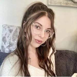 A young woman with long brown hair, glasses, and hoop earrings, sitting on a sofa in a living room with a neutral-colored wall and pillow behind her.