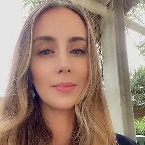 Close-up of a young woman with long wavy blonde hair and light makeup, standing outdoors near a wooden structure and trees, smiling softly.