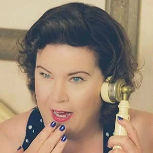 Woman with short curly dark hair talking on a vintage telephone, touching her lips with her fingers, wearing a polka dot top and displaying blue nail polish.