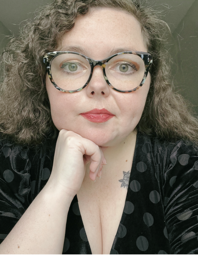 A woman with curly hair wearing large black and tortoiseshell glasses, red lipstick, and a black polka dot top, resting her chin on her hand.