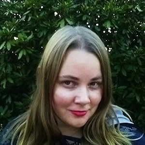 A young woman with light brown hair and fair skin smiling outdoors in front of green foliage.