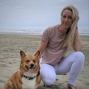 A woman with long blonde hair sitting on a sandy beach next to a tan dog with a collar.