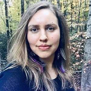 A woman with long blonde hair wearing a dark sweater stands in a wooded area with autumn foliage.