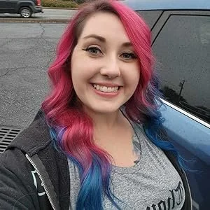 Young woman with pink and blue hair smiling outdoors near a blue vehicle.