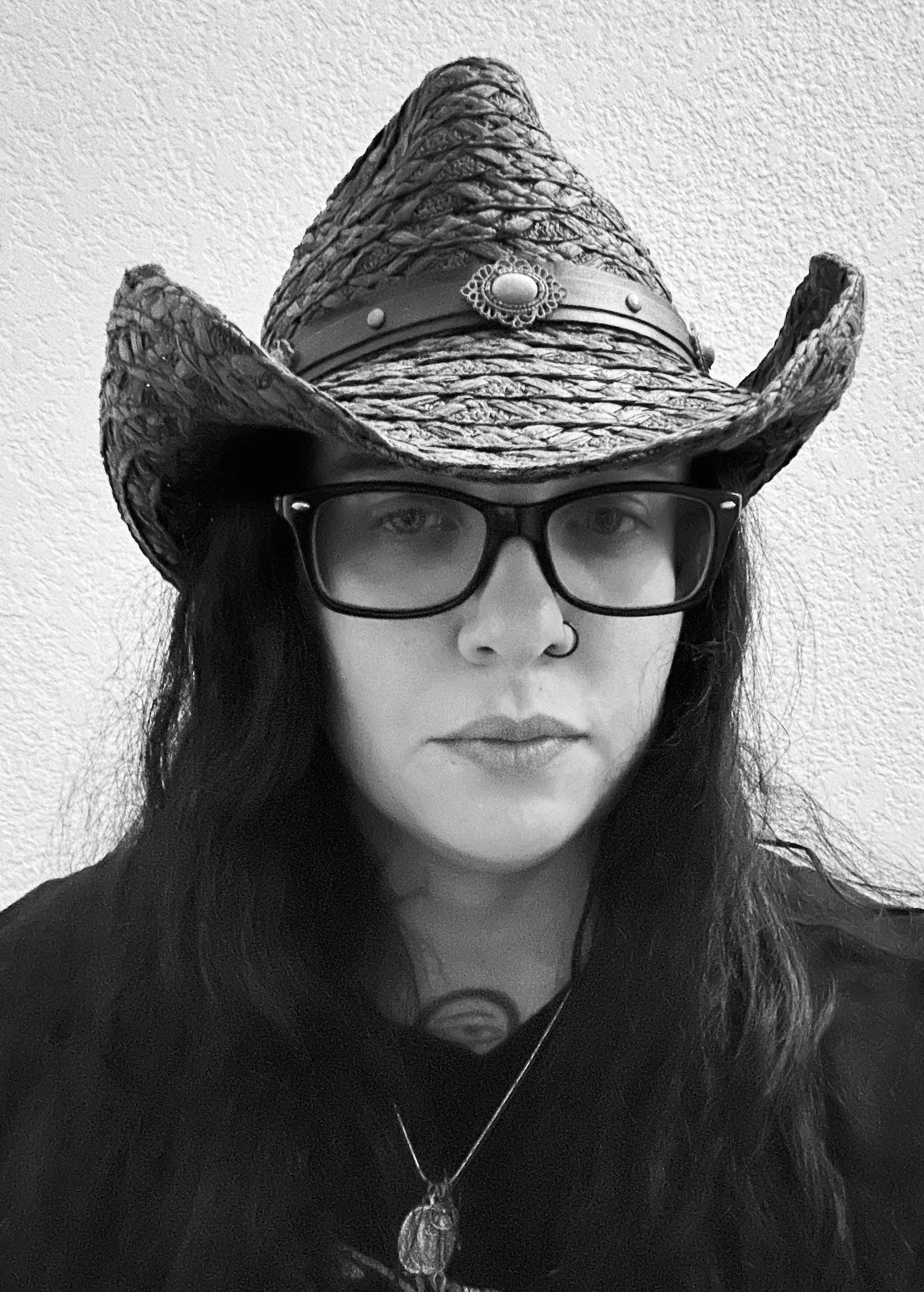 A woman wearing glasses, a nose ring, a necklace, and a cowboy hat with a decorative band, in a black and white photo.