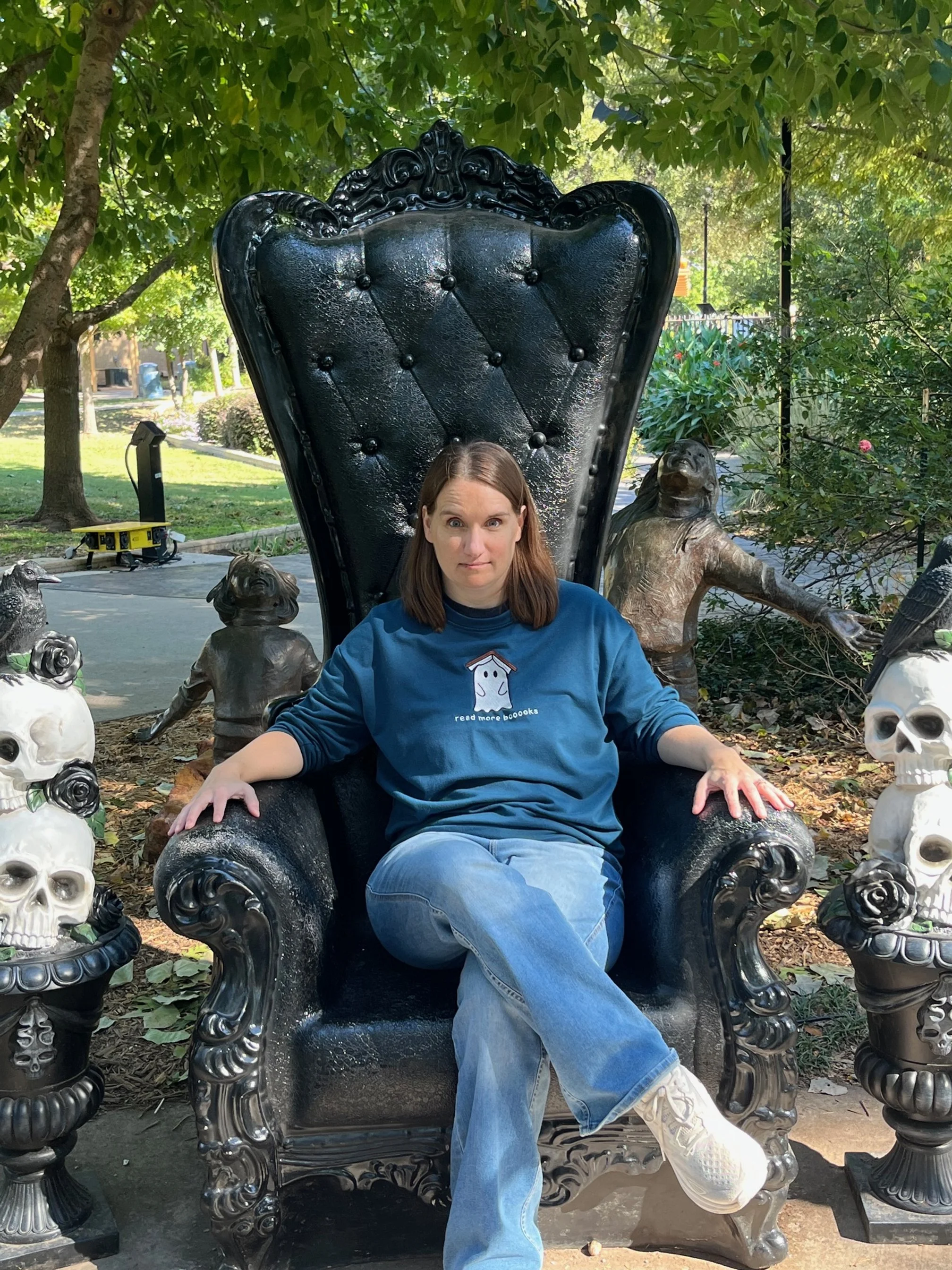 A woman sitting on a large ornate black throne-like chair outdoors, surrounded by sculptures of skulls, a dog, and a dog-like figure, with leafy trees in the background.