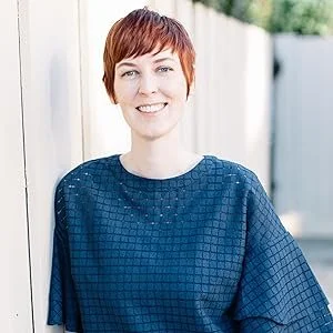 Woman with short red hair smiling outdoors, wearing a blue textured top, standing next to a white fence.