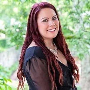 Young woman with long red hair smiling outdoors, wearing a black top and pearl necklace.