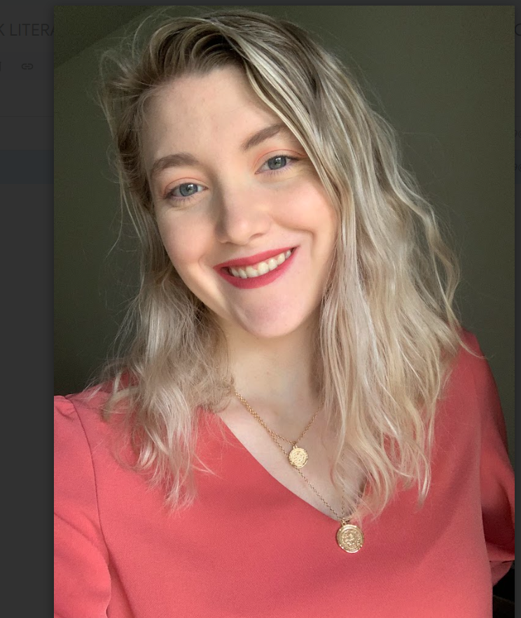 A young woman with blonde, wavy hair, wearing a pink top and gold necklaces, smiling at the camera.