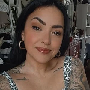 Close-up of a woman with dark hair, makeup, earrings, and tattoos, smiling in an indoor setting with furniture and decor in the background.