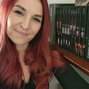 Young woman with pink hair smiling next to a bookshelf of 'Narcissa' novels.
