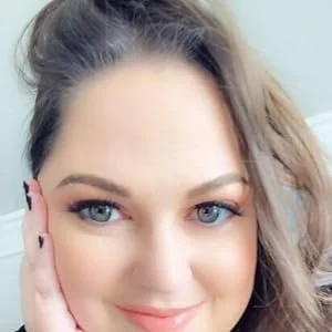 Close-up of a woman with long, wavy brown hair and blue eyes, smiling lightly, touching her face with her left hand.