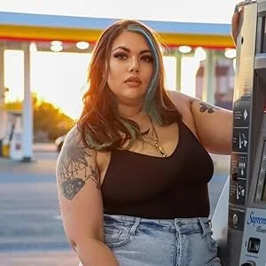 A young woman with colored hair and tattoos is standing at a gas station, wearing a black top and denim shorts, with sunlight in the background.