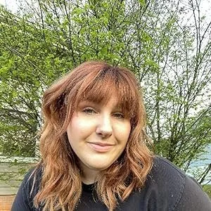 A woman with wavy, reddish-brown hair and bangs smiling outdoors with green trees in the background.