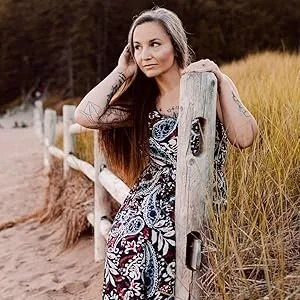 A woman with long brown hair wearing a patterned dress leaning on a wooden fence on a sandy path in a grassy field.