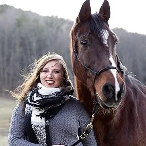 A woman smiling in a gray coat and white scarf standing next to a brown horse with a white star on its forehead, outdoors with a hill in the background.