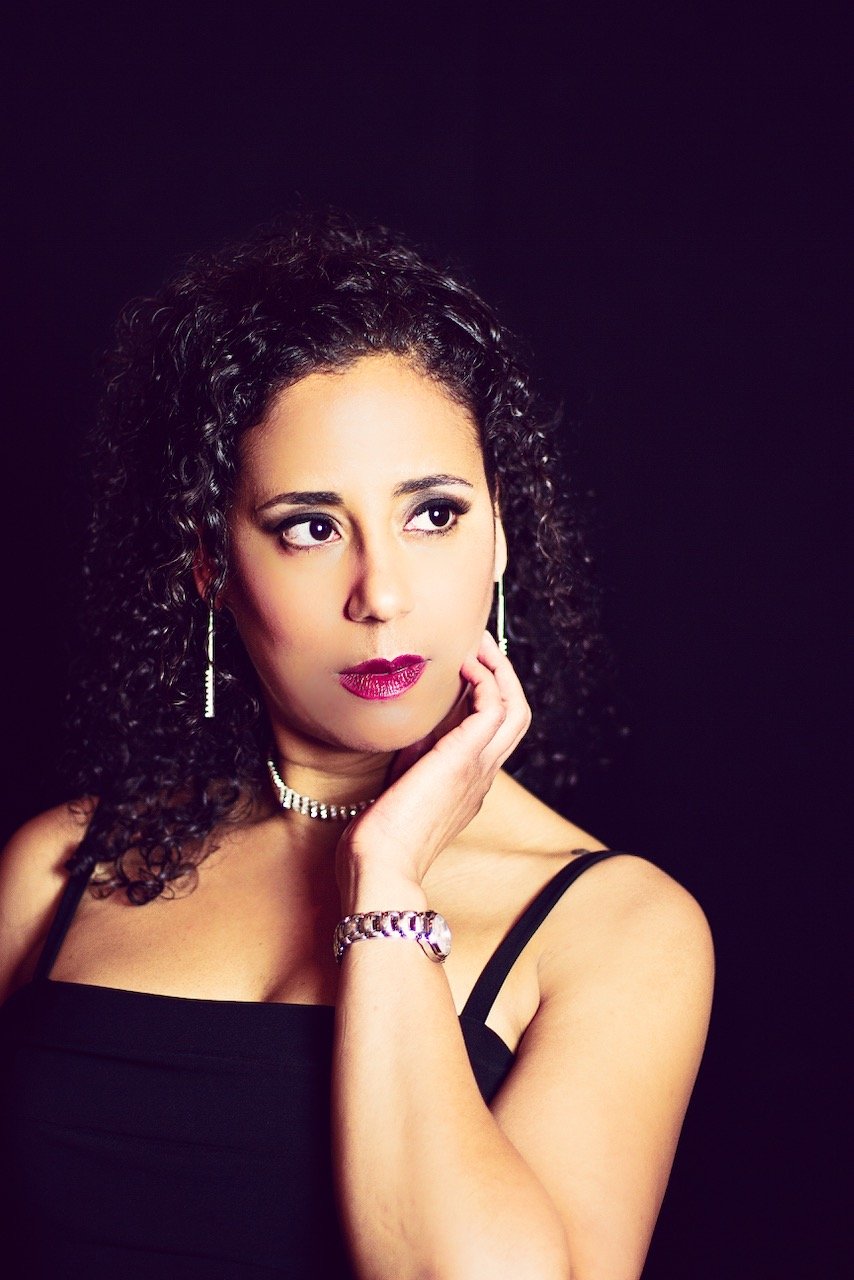 Portrait of a woman with curly dark hair, wearing a black sleeveless top, jewelry including earrings, a bracelet, a necklace, and makeup with dark lipstick, posing thoughtfully against a black background.