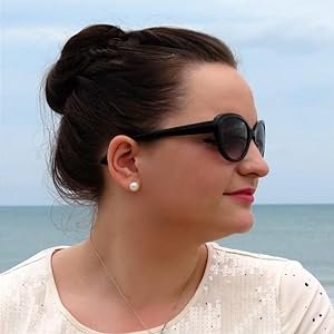 Profile of a woman with dark hair in a bun wearing black sunglasses and pearl earrings, against a cloudy sky and ocean background.