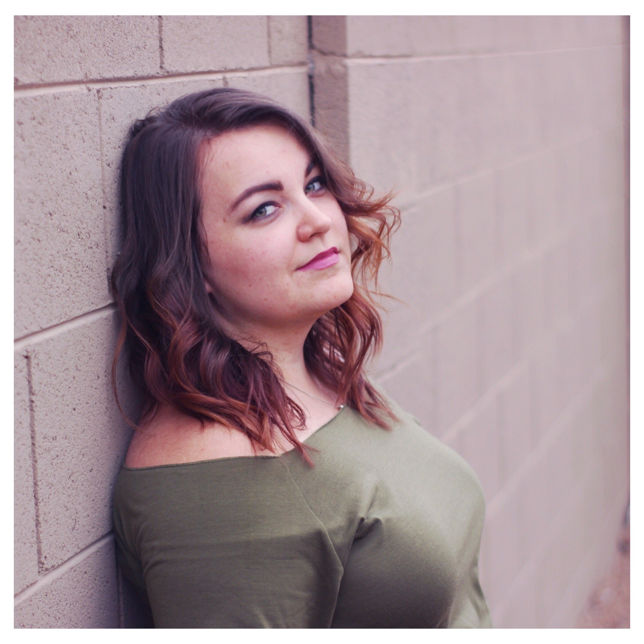 A woman with wavy, reddish-brown hair and light skin leaning against a brick wall, looking at the camera with a slight smile. She is wearing an off-the-shoulder olive green top.