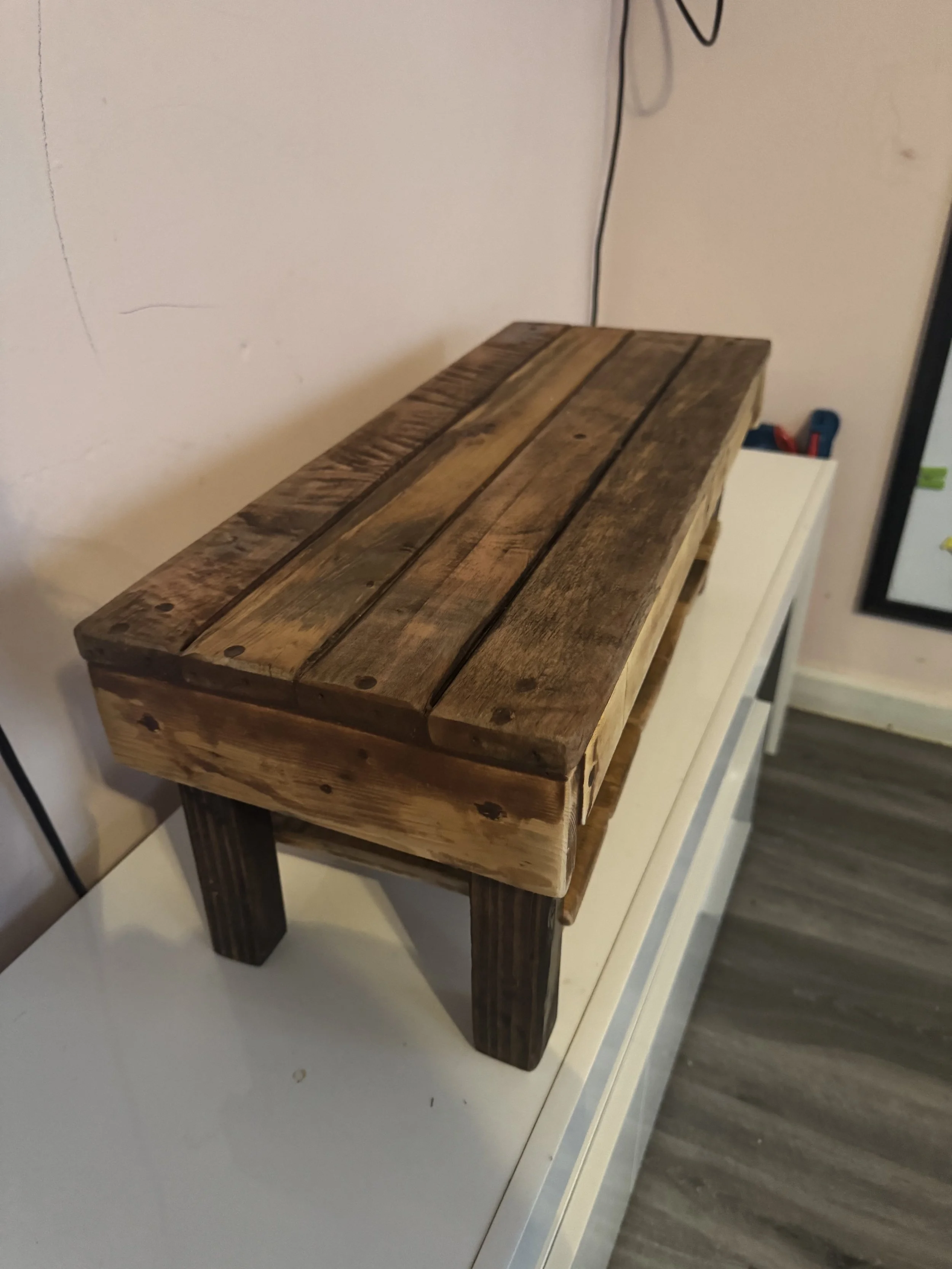 Small wooden table made of reclaimed wood with three legs, placed on a white surface against a wall.