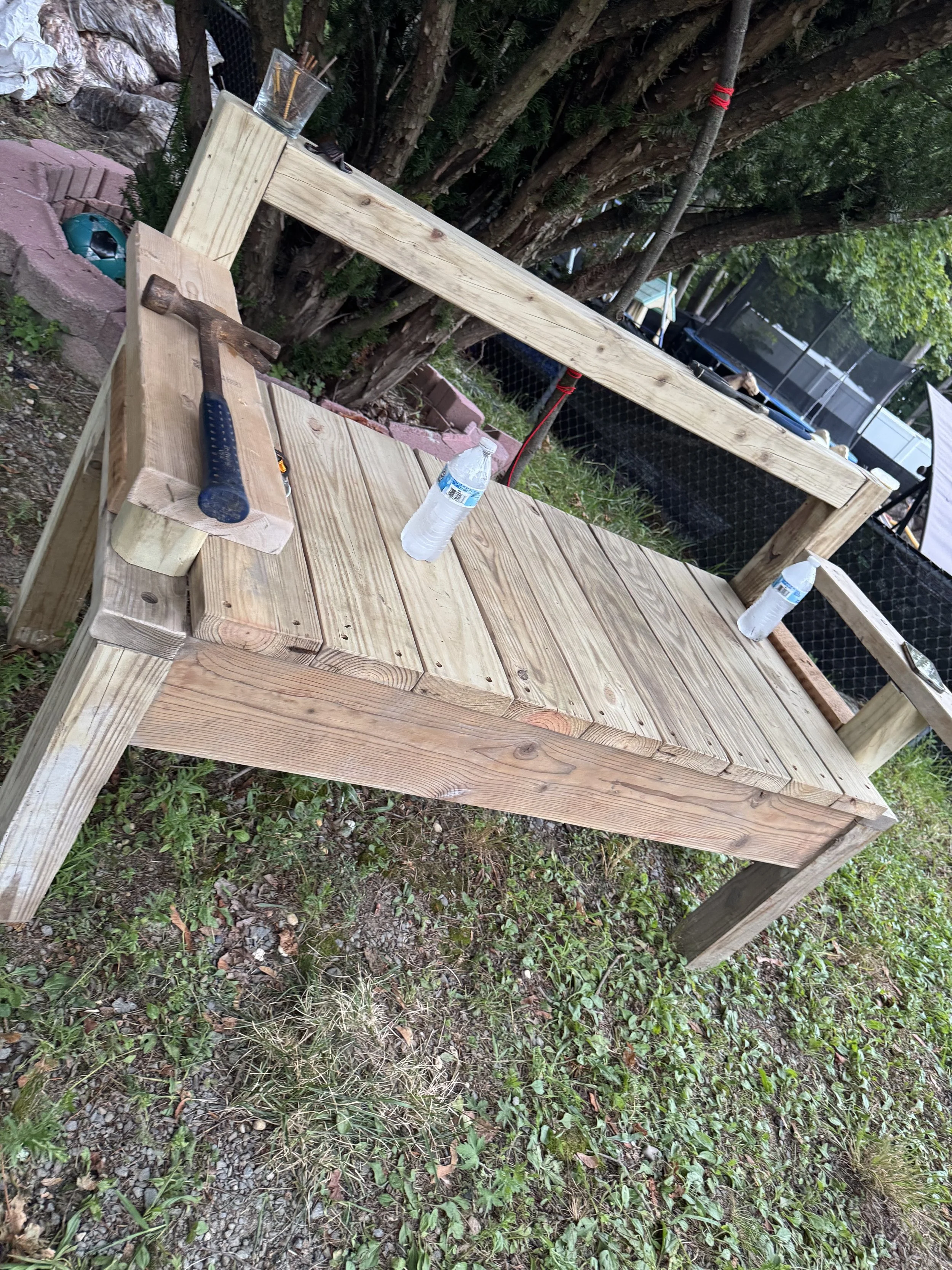 Unfinished wooden outdoor bench under trees.