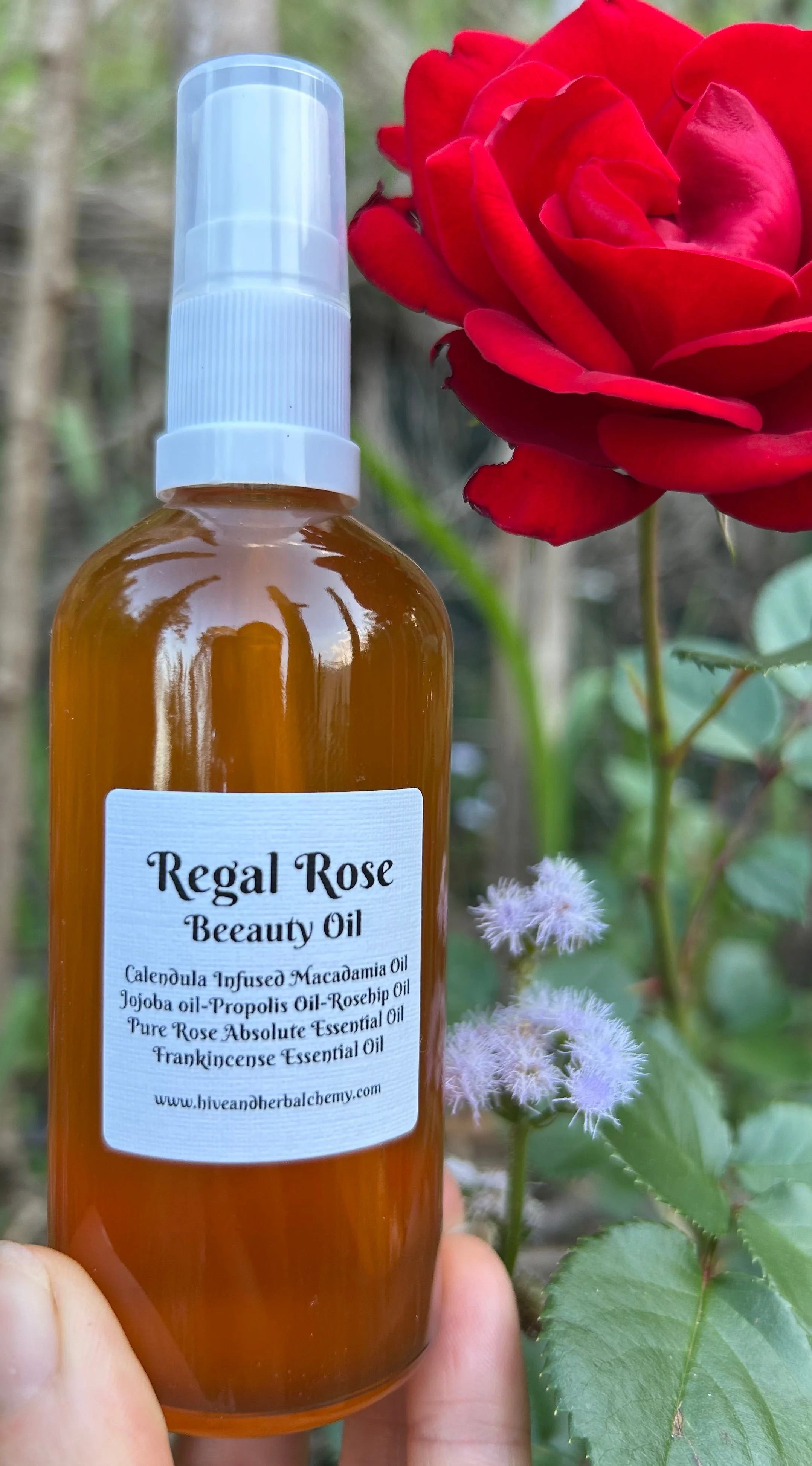 Regal Rose Beeauty Oil spray bottle held up beside a red rose in a garden